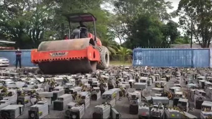 A steamroller labeled "HAMM" (visible on its side) crushes a massive collection of cryptocurrency mining rigs (ASICs) arranged in rows, illustrating the post's point about Bitcoin mining operations shutting down as the industry becomes unprofitable—and the resulting liquidation of mining equipment.