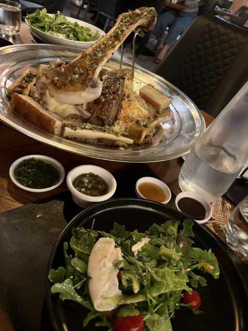 A restaurant meal featuring a large meat and bread bone marrow dish in a silver platter, accompanied by small bowls of sauces, a green salad with tomatoes in a black bowl, and a bottle of water.