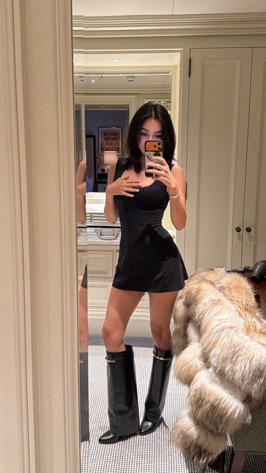 A woman takes a mirror selfie in an elegant black mini dress paired with tall black knee-high boots, standing in a sophisticated dressing room with a furry garment draped nearby, fitting the "Gm Uptober or Rektober?" post about crypto market uncertainty by showcasing a confident, stylish mirror selfie aesthetic.
