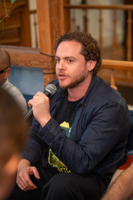 A man with curly brown hair and a beard speaks into a microphone while wearing a dark blue button-up shirt over a graphic t-shirt, seated in what appears to be a panel discussion.