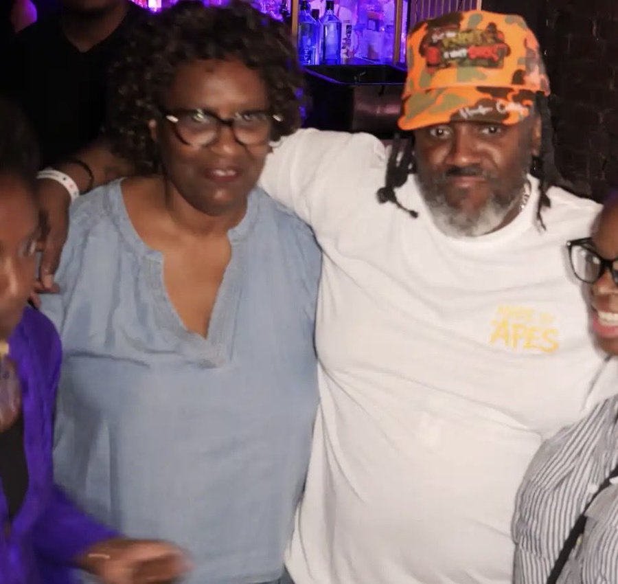 A man wearing a white 'Bored Ape' t-shirt and an orange camouflage cap poses with his arm around a smiling woman in glasses and a blue denim top.
