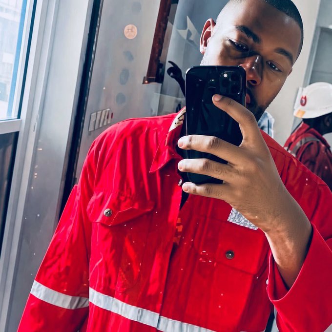 A selfie photograph of a man wearing a red work shirt with reflective stripes, holding up a black smartphone, with another person partially visible in the background.