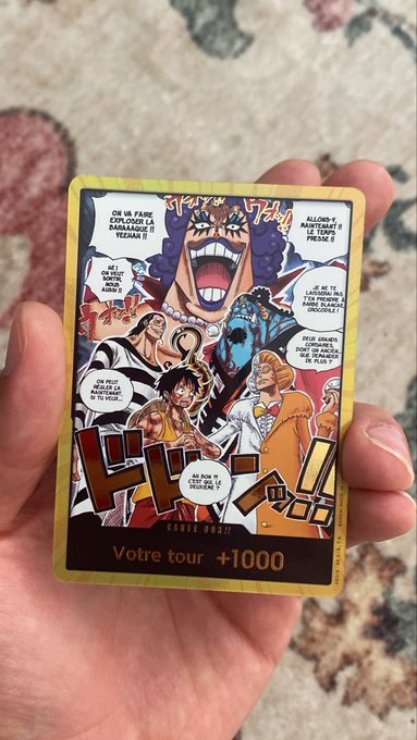 A hand holds a colorful Japanese trading card from a manga (appears to be One Piece), labeled 'Carte Robe' with the text 'Votre tour' and '+1000' visible at the bottom.