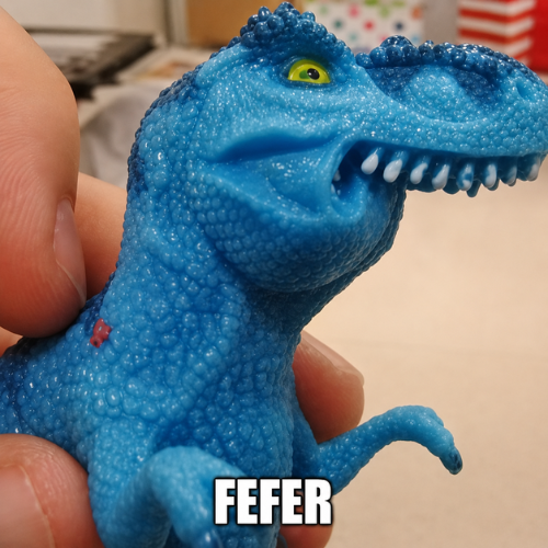 A person's hand holding a small blue plastic T-Rex dinosaur toy with green eyes and white teeth, labeled 'FEFER' in white text at the bottom.