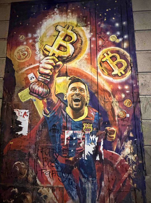 A street mural depicting a soccer player (appearing to be Messi) holding up a Bitcoin symbol, surrounded by other Bitcoin logos and stylized artwork.