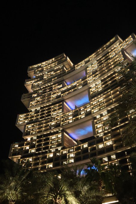 A towering, illuminated hotel high-rise at night showcases its curved, terraced architecture glowing warmly against the dark sky, with palm trees framing the foreground. The building's distinctive stacked, cantilevered balconies lit up serve as a visual representation of "GN," likely referencing a luxury resort or notable hospitality brand associated with the image.