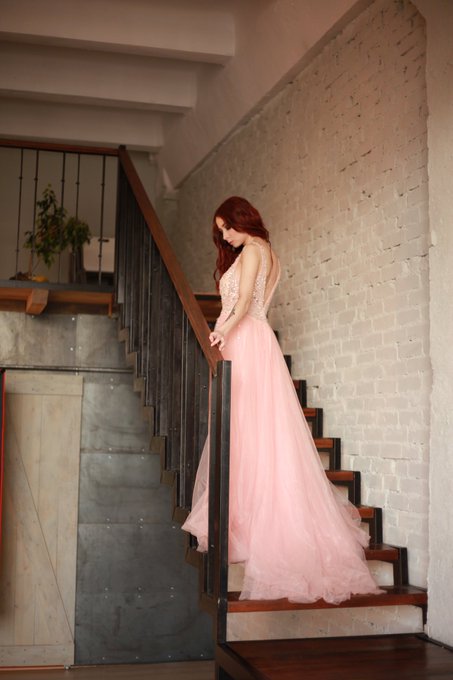 A red-haired woman in a flowing pale-pink formal gown stands on a wooden-and-metal staircase beside a white brick wall.