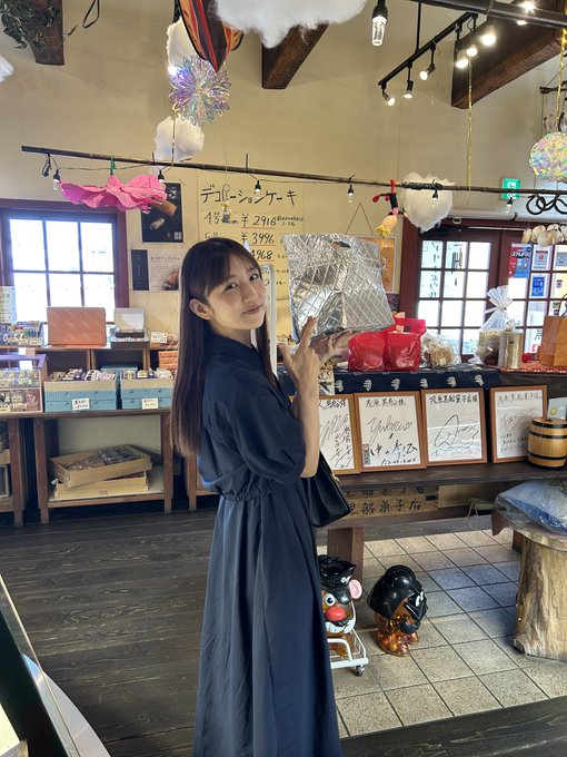 A woman in a dark blue dress holds up a silver foil-wrapped prototype snack package inside a rustic shop filled with Japanese merchandise, where signage reads "デコポーシケーキ" (Deco Sea Cake) priced at ¥294 and ¥399. The image visually connects to the caption by showing Yuuko-rin proudly displaying the still-unfinished prototype treat she received during her visit.