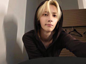 A selfie of a young man with blonde hair wearing a black hoodie, posed in what appears to be a dressing room with hangers visible in the background. The hashtags reference "AEN," "JUNSEO," and nicknames associated with the K-pop group, suggesting this is a personal photo shared by the artist.