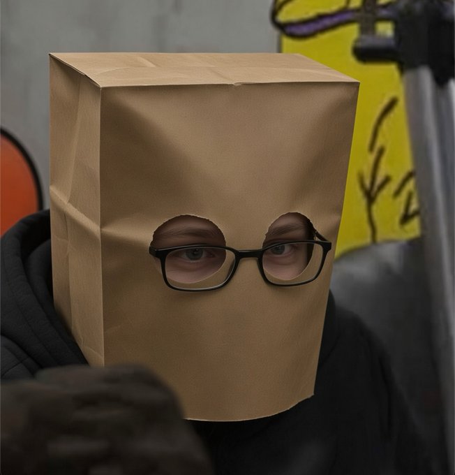 A person wearing a black hoodie with a brown paper bag over their head, with eye holes cut out to show their eyes and black-rimmed glasses, standing in front of graffiti.