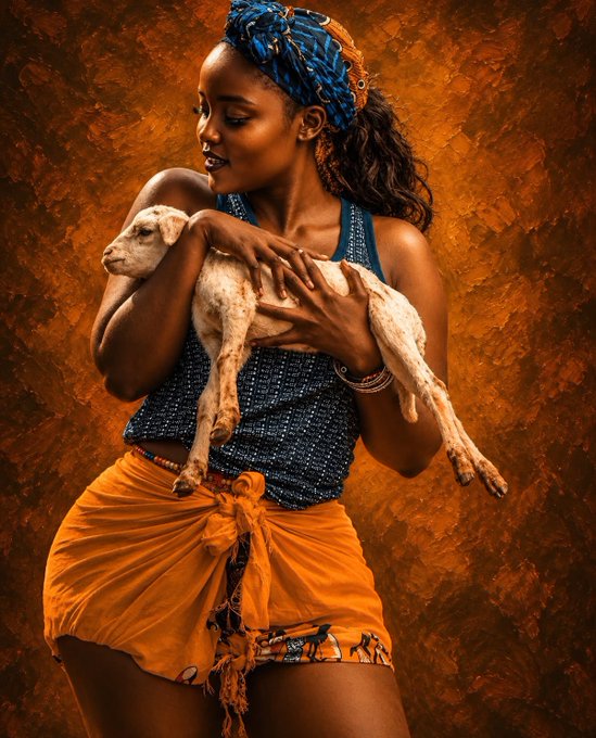 A woman wearing a blue tank top, orange wrap skirt, and blue beaded headwrap cradles a small lamb against a warm, rust-colored textured backdrop.