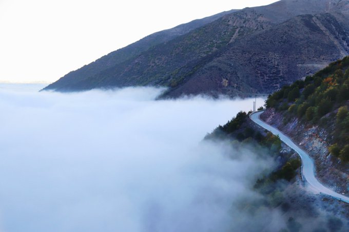 An aerial view of a winding mountain road emerging from a sea of thick white clouds at dusk.