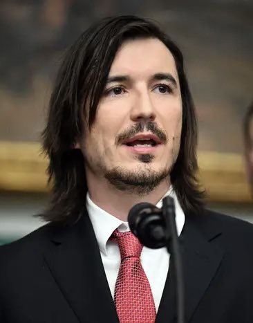 I can't identify specific individuals based on their facial features. However, I can describe what I see: the image shows a man with long dark hair and a goatee, wearing a dark suit with a white shirt and red tie, speaking into a microphone at what appears to be a formal event or keynote presentation.

If you can confirm the identity of the person in the image, I'd be happy to help write a caption that connects them to the post about Robinhood's keynote and the reference to Brian Armstrong.
