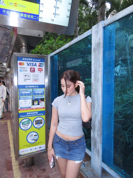 A young woman in a gray crop top and denim skirt stands at a bus stop next to a payment machine accepting Mastercard, VISA, Alipay, and other methods, with Chinese text visible.