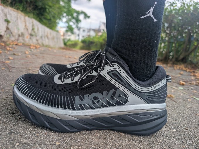 The image features a person wearing black and gray sneakers, with their feet positioned on a sidewalk or pavement. The shoes are described as "NOKA" style, which suggests that they might be a popular brand among sneaker enthusiasts. The person is also wearing socks, adding an extra layer of detail to the overall scene.