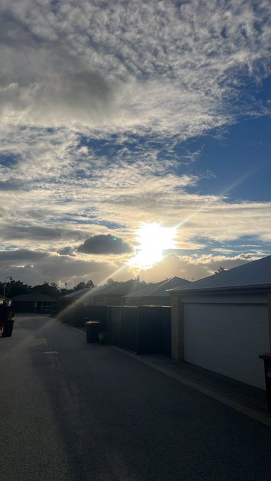 A suburban street scene at sunset or sunrise, with bright sunlight breaking through scattered clouds above rows of houses with white garage doors.