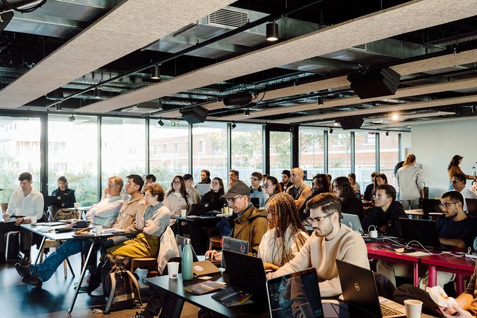 A diverse group of people sits in a modern, well-lit coworking space with laptops and monitors during a session or meeting.