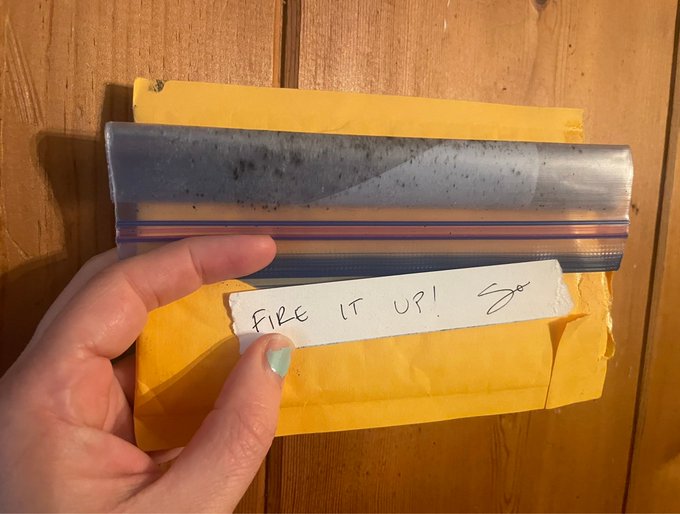 A hand holding a yellow padded envelope with a note reading 'Fix it up!' written on it, with a metal ruler and paper visible on a wooden table.