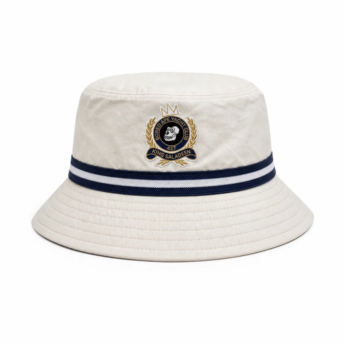 A cream-colored bucket hat with navy blue and white striped band, featuring an embroidered 'Monkey Tape Yacht Club 427 KING SALADEEN' crest with a gorilla logo.