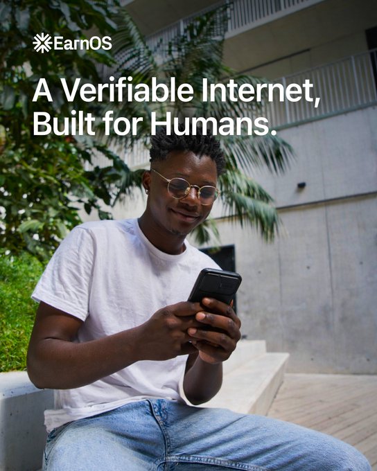 A young man wearing a white t-shirt and glasses smiles while looking at his smartphone, seated outdoors near lush greenery and a modern building. The image features the EarnOS logo and the text "A Verifiable Internet, Built for Humans," visually reinforcing the post's message about distinguishing real human activity from bots online.