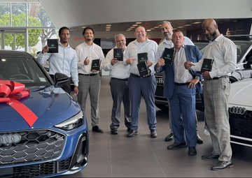 A group of men, including ZBCN/Zebec staff, pose together at a Palm Beach car dealership holding boxed prizes in front of a red-bowed Audi, celebrating a 4th of July giveaway promotion tied to the Zebec real-time payroll ecosystem.