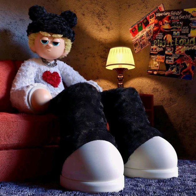 A grumpy plush doll wearing a black beanie, white heart sweater, and black furry pants lounges on a red couch beside a lamp, with posters reading "DOLL GAME FOR US? WE'VE GOT IT!" and "Play loud" on the wall behind it. The image captures the edgy, rebellious aesthetic referenced in the post teasing the upcoming "Robinhood" alpha for @dollclubnft, giving the doll NFT project a distinctly "don't sleep on this" vibe.