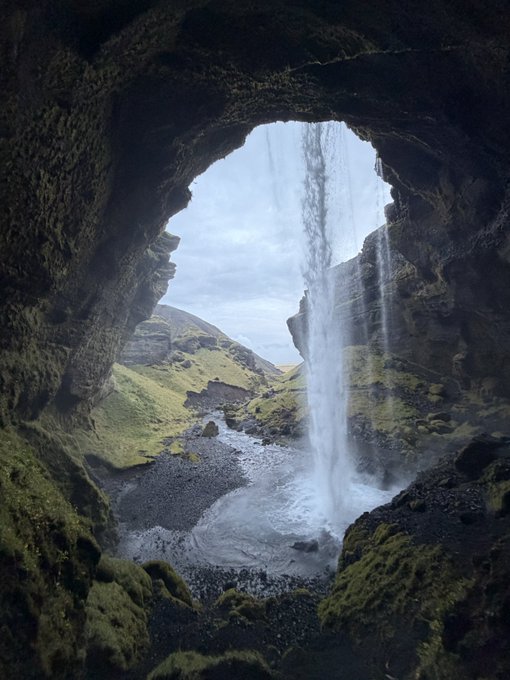 A scenic view from inside a moss-covered cave looking out at a cascading waterfall in a lush, rocky landscape.