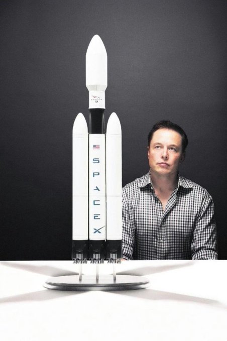 The image features a man sitting at a table with a model of a rocket ship, which appears to be made out of cardboard or foam board. The man seems to be looking at the rocket ship, possibly admiring its design or contemplating its purpose. The photo is taken from an overhead perspective and has a black background that contrasts with the white rocket ship model. The caption on the image reads "SpaceX 5muNNyusv1FNkPp7Gv3mbT3TqUzZ1EQndqLi8wnTQEY6".