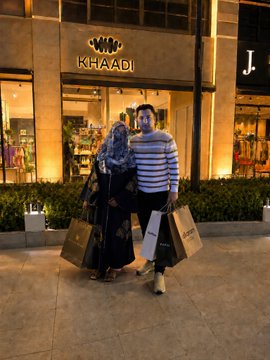 A couple stands together in front of a Khaadi storefront at night, holding shopping bags from the boutique, appearing to have just enjoyed a shopping trip. The image relates to the post as the person showcases their recent Khaadi shopping purchases before announcing they will tip $100 to three Stake users.