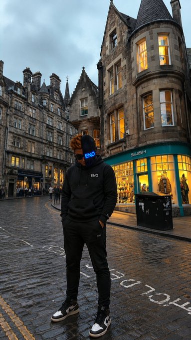 A person wearing an "ENO" branded hoodie and cap standing on a cobblestone street in what appears to be Edinburgh, with historic Scottish architecture and a "SLUSH" shop in the background.