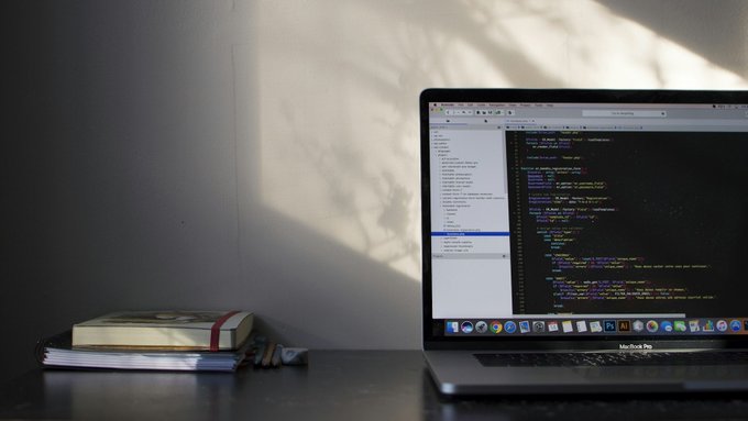A MacBook Pro displays a code editor on its screen, positioned next to a stack of notebooks on a desk, with sunlight casting shadows on the wall behind—illustrating a coding or development workspace that complements the post's suggestion of integrating CoinGecko with ClashOfAI.