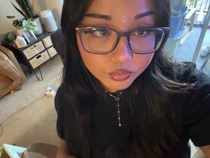 A selfie of a young woman with dark hair, glasses, a nose ring, and a black top, sitting indoors with a plant-filled shelf and sliding glass door visible in the background.