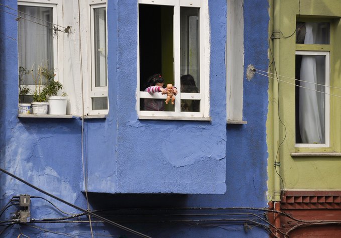 A blue building features an open window where a person holds a teddy bear, with another dark-haired person visible nearby, while potted plants sit on a windowsill and tangled wires run along the weathered exterior.