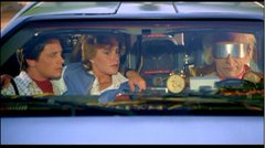 Two men inside a car at dusk, one in a blue suit holding a flashlight, appearing to be a film still.