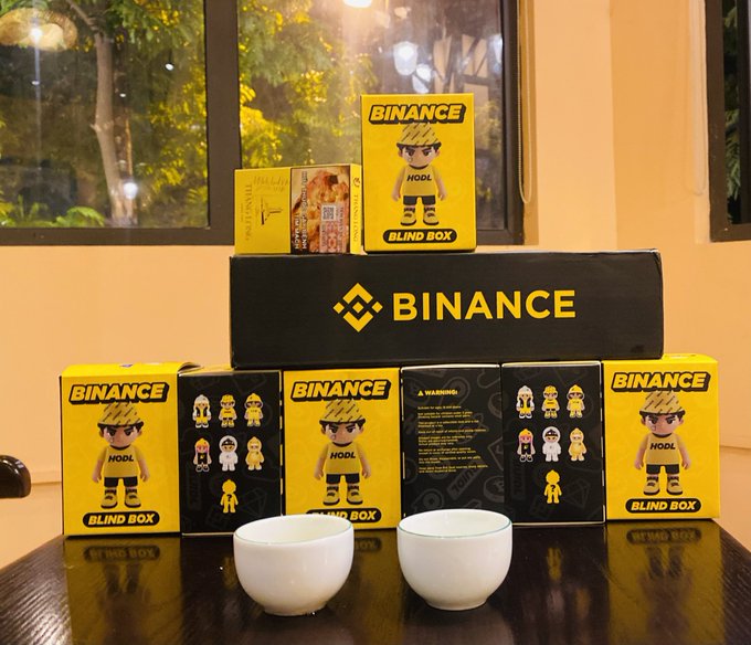 A display of yellow and black "BINANCE" "BLIND BOX" merchandise featuring a character labeled "HOOL" in a yellow shirt, arranged on a table with two white teacups.