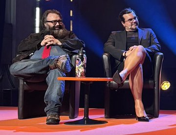 Two men sit in black leather chairs on a stage with a small red table between them, appearing to be part of a panel discussion or talk show.