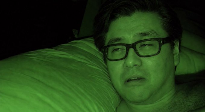 A close-up shot, likely taken with night vision, shows a man with glasses lying in bed with his eyes half-open.