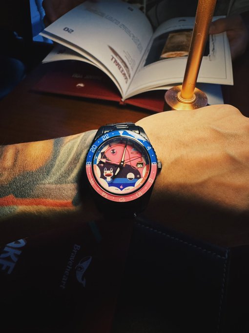 A tattooed arm wearing a GMT-style watch with a red and blue bezel and a distinctive dial sits next to an open magazine on a dark wooden table under warm lighting.