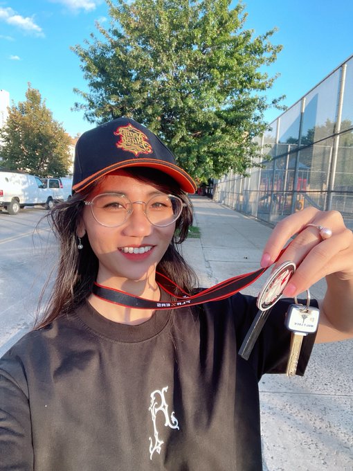 A young woman wearing glasses and a black cap smiles while holding up a set of keys, likely marking a new home or property.