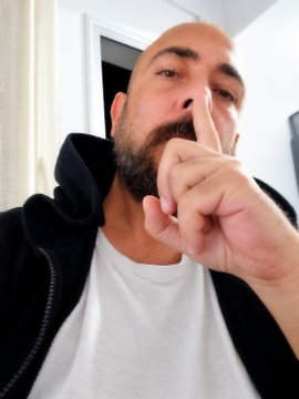 A bearded man wearing a black hoodie over a white t-shirt is captured in a close-up selfie, holding his index finger up to his nose in a "shushing" gesture.