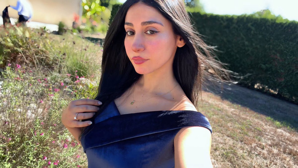 A young woman with long dark hair wearing a navy blue off-the-shoulder dress and a delicate gold necklace poses outdoors with greenery and pink flowers in the background.