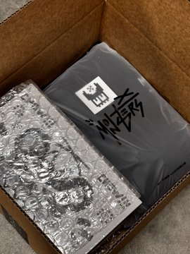An open cardboard shipping box containing items wrapped in black plastic and silver bubble wrap, with handwritten text on the black wrapping.
