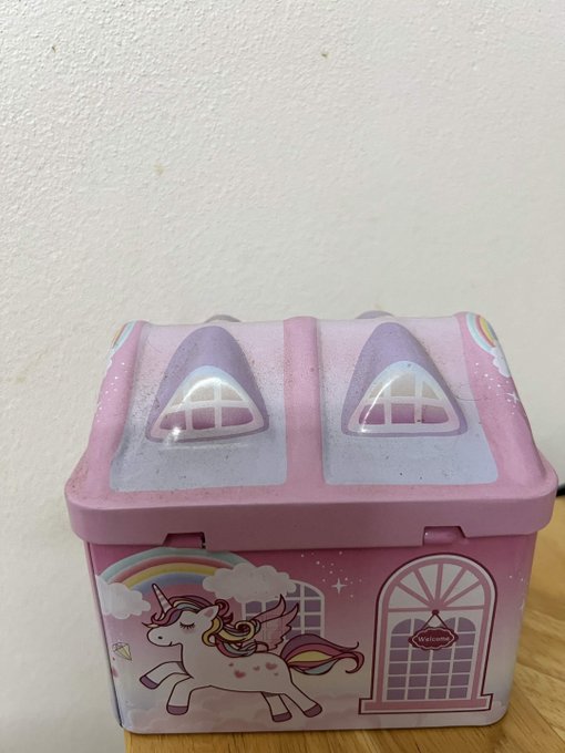 A pink plastic children's storage box decorated with a unicorn, rainbow, clouds, and a castle design, featuring two dome-shaped openings on the lid.