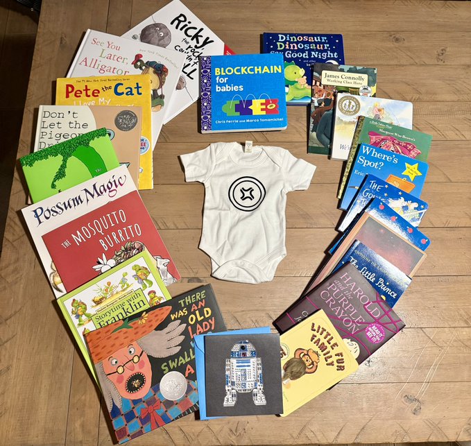 A white baby onesie with a black geometric logo sits in the center of a heart-shaped arrangement of children's books, including titles like "Pete the Cat," "Ricky," "Possum Magic," "Blockchain for babies," and "The Mosquito Burrito."