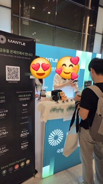 A person wearing a backpack stands at a trade show booth featuring the 'MANTLE' cryptocurrency logo and displays with emoji faces obscuring part of the view.