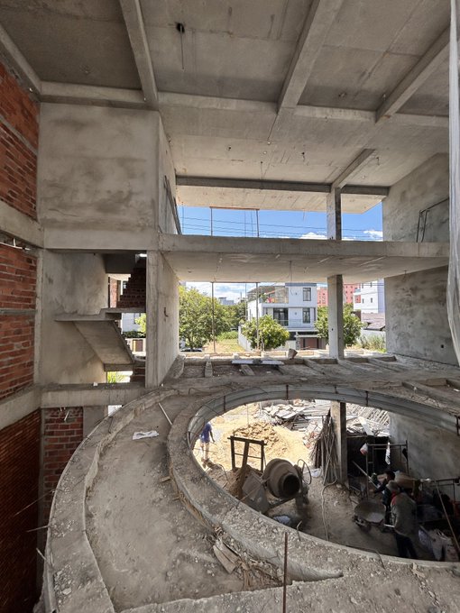 A view from inside a building under construction, showing an unfinished concrete spiral staircase in the foreground and a cement mixer visible through a circular opening, with residential buildings in the background.