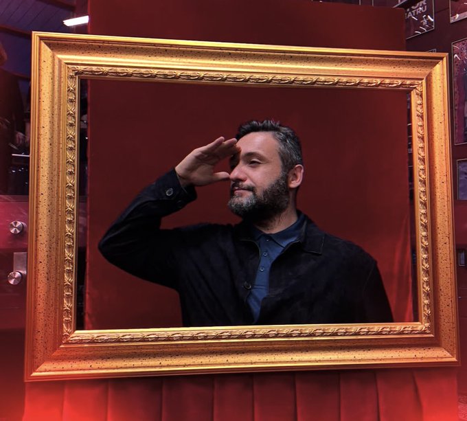 A man with a beard, wearing a dark shirt, posing inside a large ornate gold picture frame against a red background with his hand near his forehead.