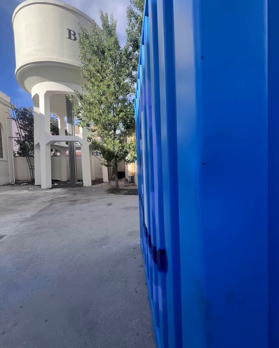 The image features a large blue water tower with a tree growing beside it, as well as a blue portable toilet or porta potty nearby. The water tower has the words "Bran" written on its side, which could be related to the city of Lisbon. The photo is taken from an angle that emphasizes the size and height of the water tower while also showcasing the tree growing next to it.