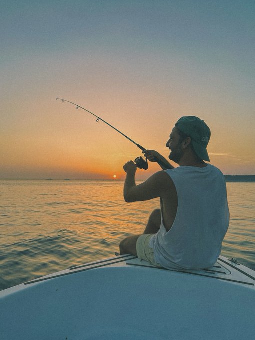 A man in a white tank top and backwards cap sits on the edge of a boat, fishing with a rod silhouetted against a serene orange sunset over the water.