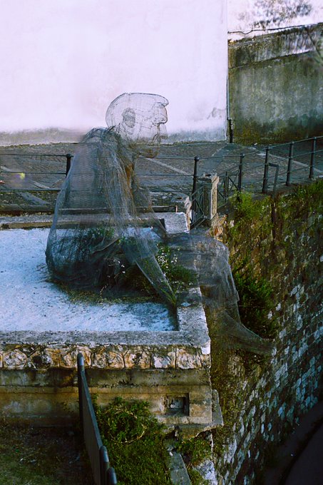 A wire mesh sculpture of a seated figure sits on an old stone balcony overlooking a grassy area with a railing, rendered with a soft, slightly blurred effect.